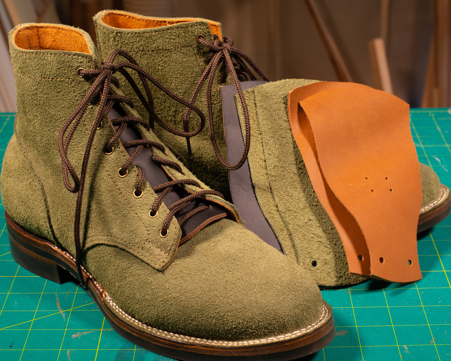 A pair of green kilties (false tongues) for boots displayed on a table with a pair of boots and a piece of brown leather.