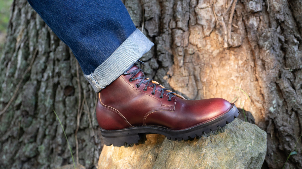 A close-up of a person's foot wearing a Service Boot in Horween Color 8 Chromexcel, standing on a rock against a textured tree trunk background.